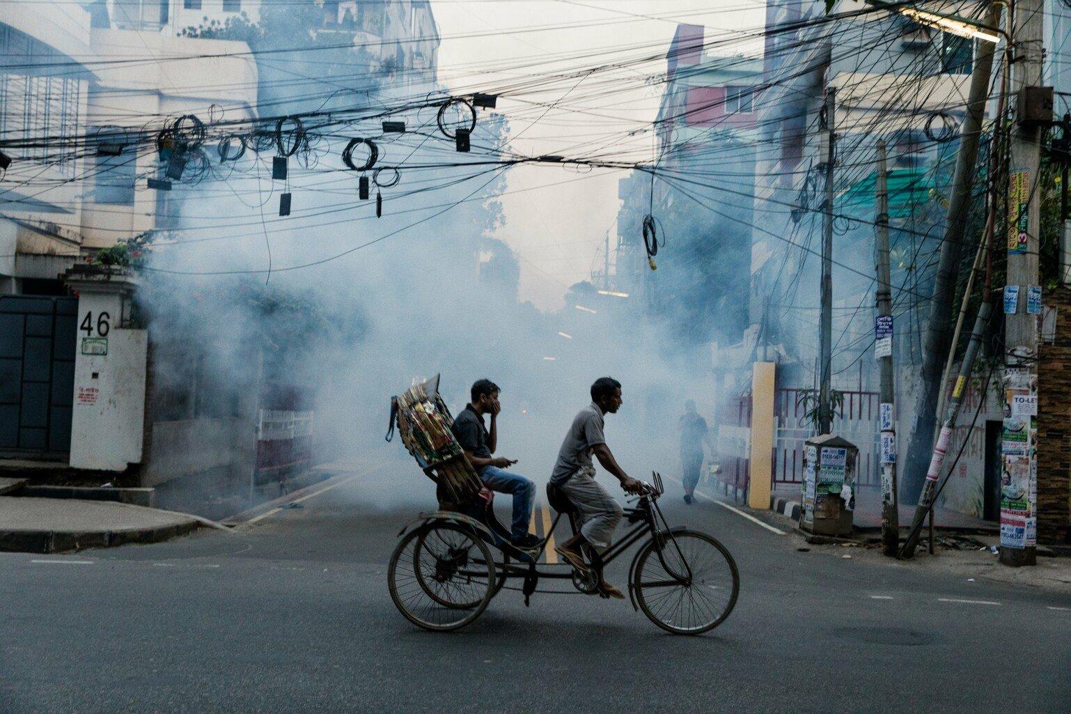 Two people cycling on a street in Uttara which is being sprayed with insecticide to kill mosquitoes. Zoonotic diseases are a major global public health problem.