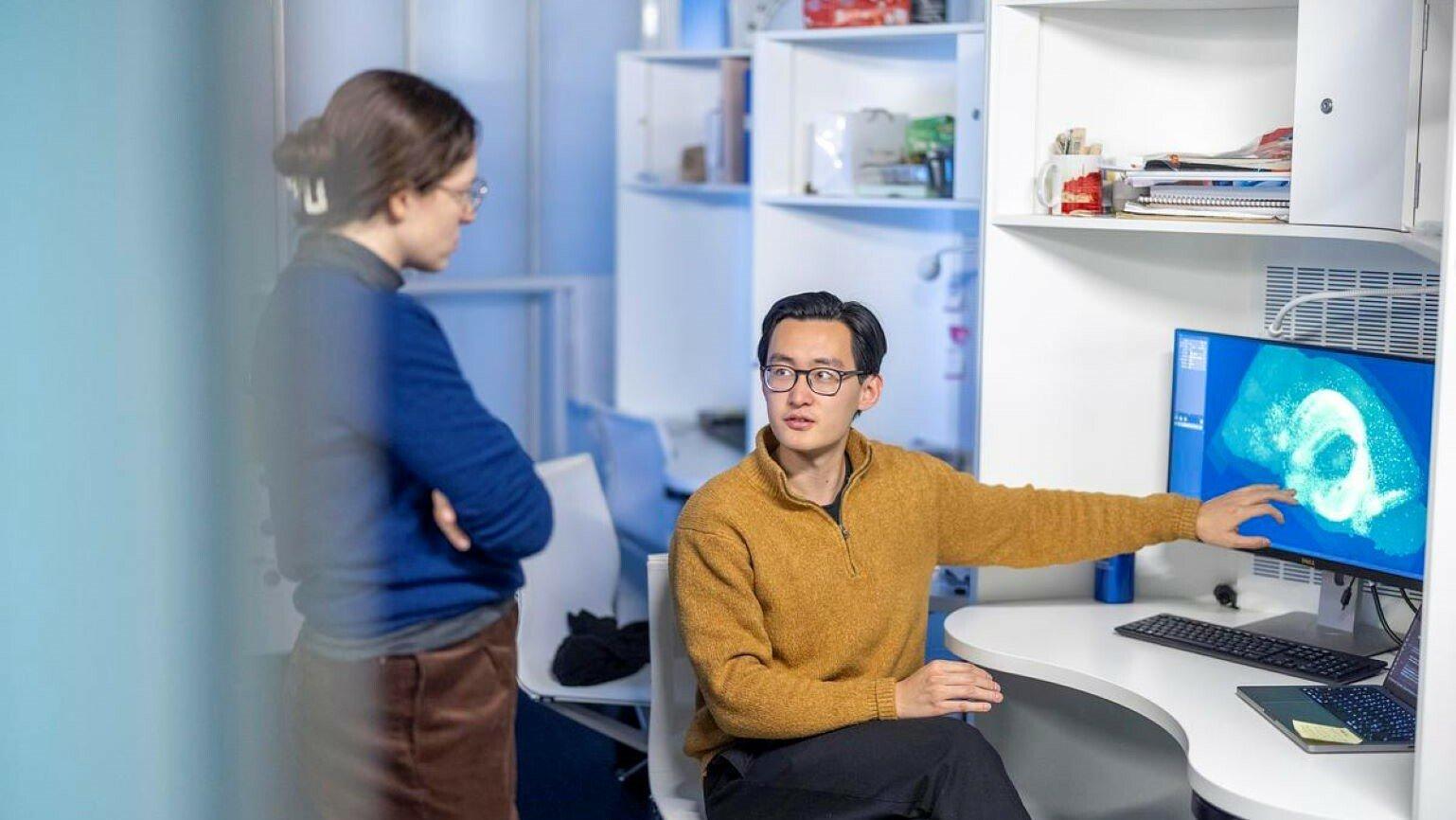 A woman in a blue jumper listens intently to a man in a yellow sweater pointing to a computer monitor.