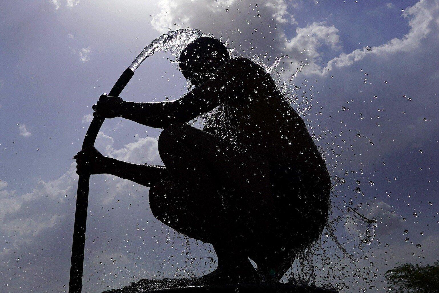 A man is silhouetted against the sun as he bathes using a water hose on a hot day.