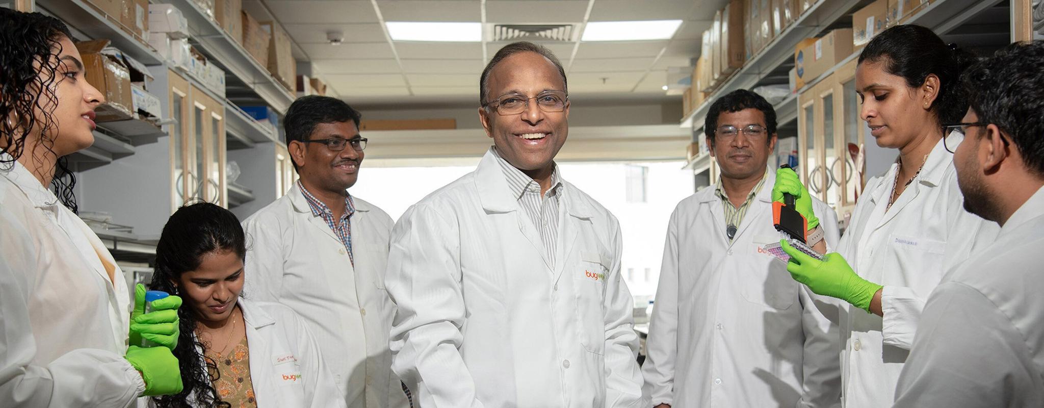 Dr Anand Anandkumar stands in the middle of his lab, smiling at the camera. He is surrounded by six colleagues – everyone is wearing a white labcoat, and several are wearing bright green latex gloves as they work pipetting into test trays.