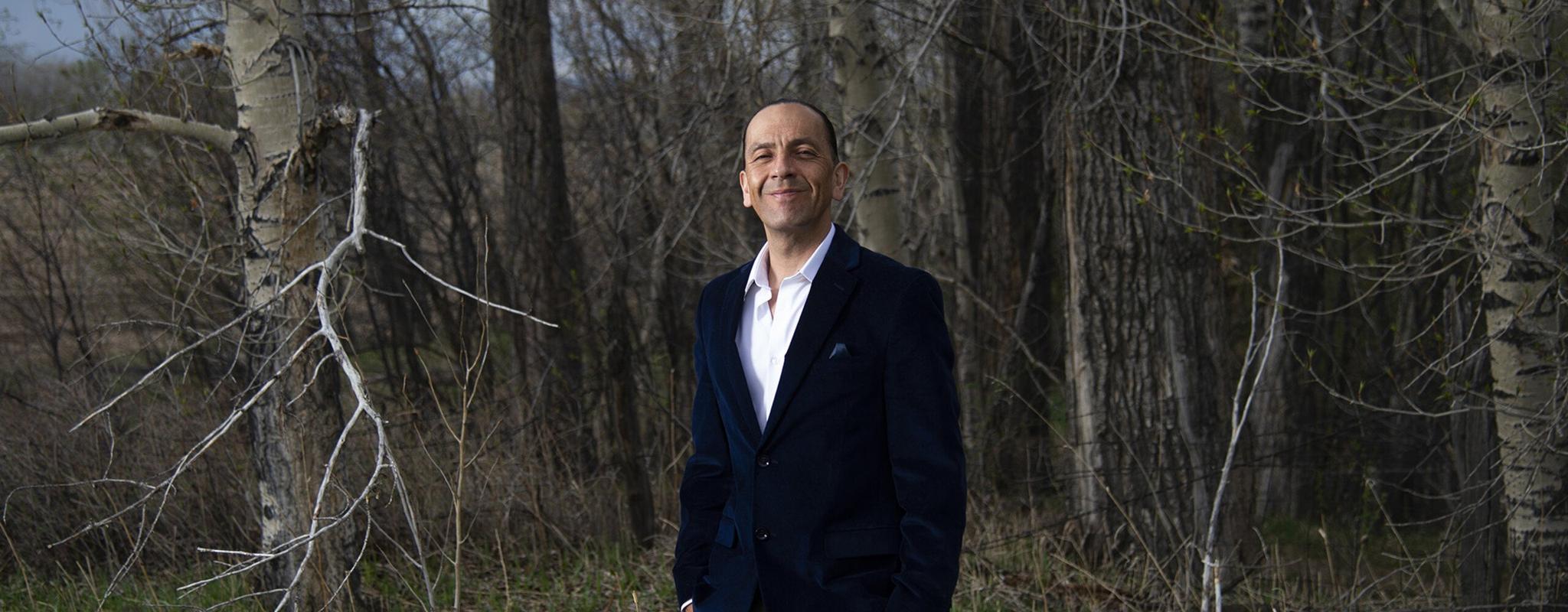 Shane Doyle stands proudly in a grassy forest. He's wearing smart grey suit trousers, a dark blue velvet blazer and a matching pocket square. His hands are in his pockets and he's smiling at the camera.