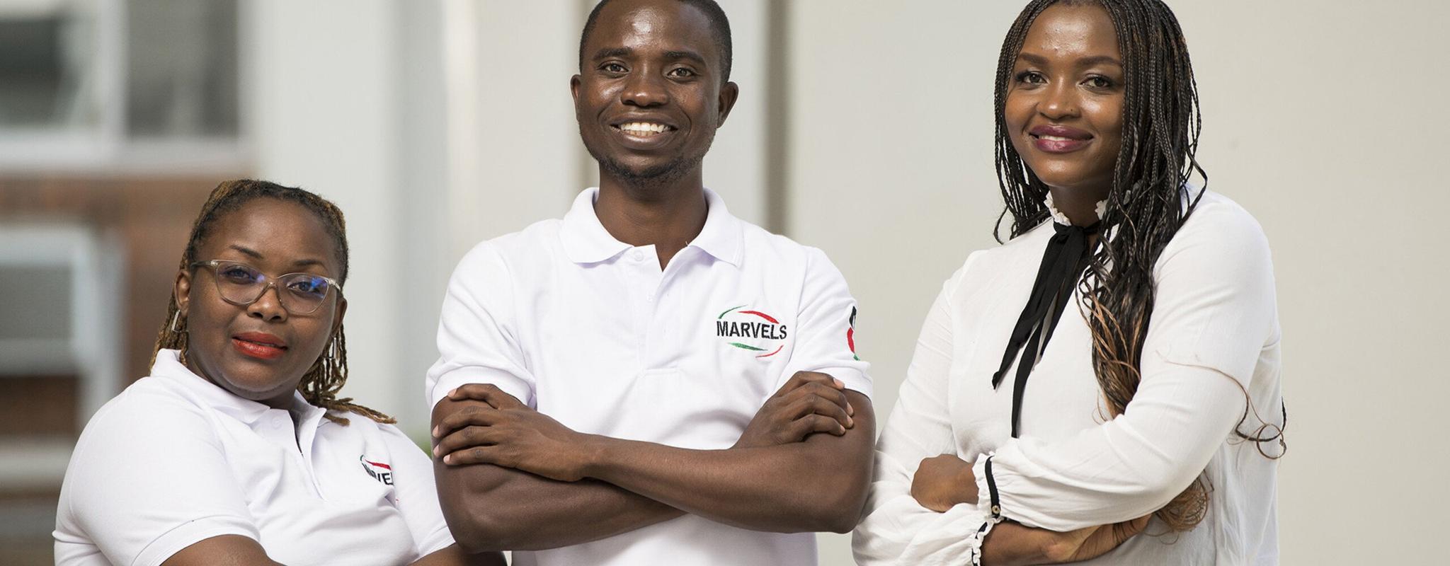 Dr Dingase Dula, a young black woman, stands smiling with her arms crossed next to two of the volunteers from her research study. The volunteers are wearing crisp white polo shirts branded with the word 'Marvels', and are also smiling with their arms crossed.
