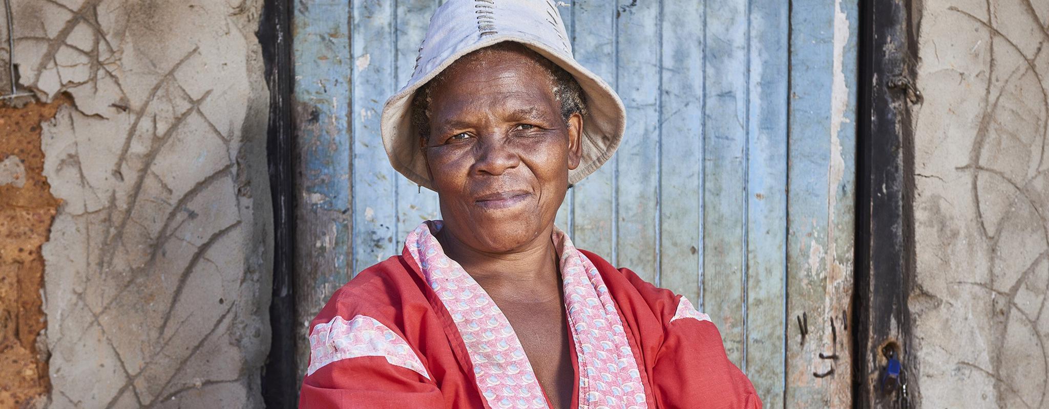 Makita Magwaza stands firmly, arms crossed, in front of a faded blue door. She wears a colourful red dress and smiles from beneath her white bucket hat.