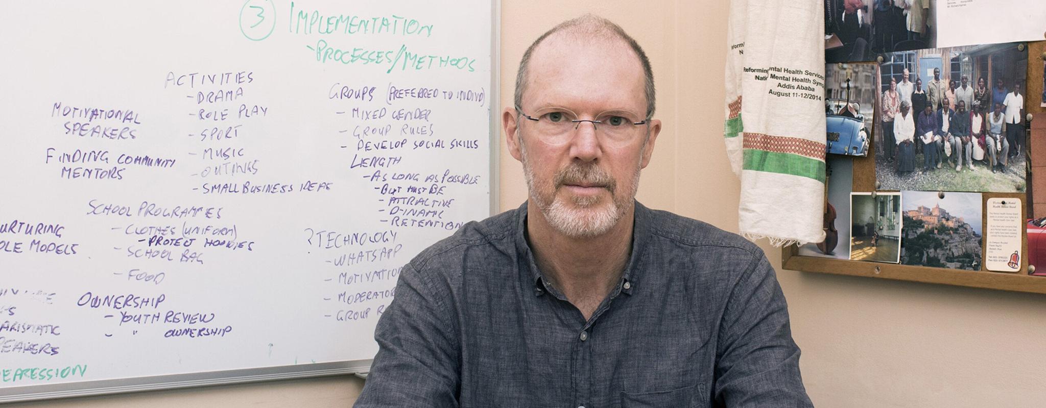 Professor Crick Lund sits at a table in the corner of an office wearing a dark linen shirt and frameless glasses. Behind him, a whiteboard is covered in scrawled notes and a pinboard displays clippings from newspapers, team photos and a memorial scarf from a conference in Addis Ababa.