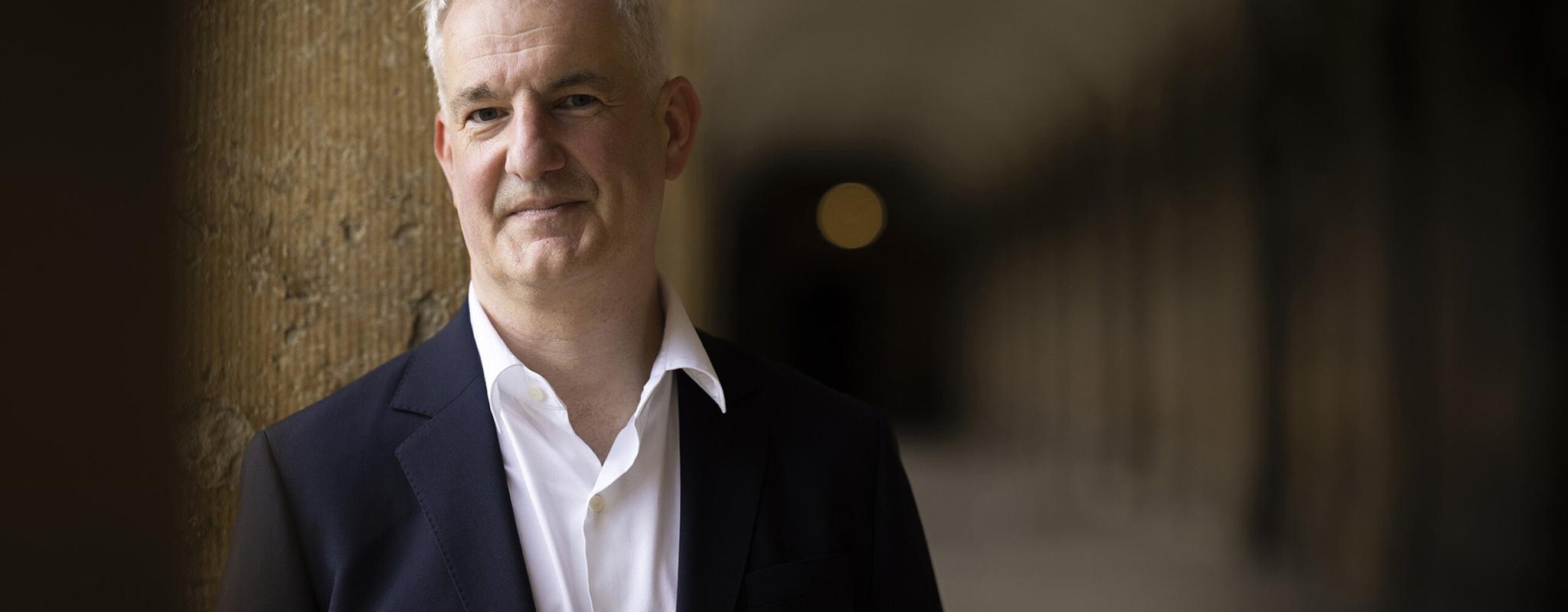 A middle aged man with white hair, a blazer and open collar shirt, stands leaning on a pillar in one of the Oxford University's grand stone hallways.