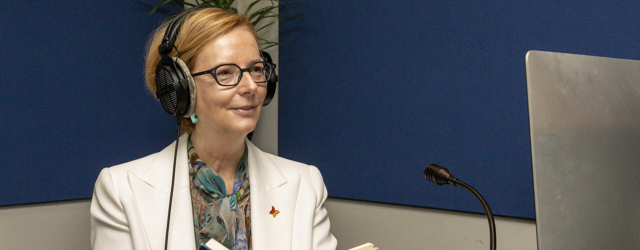 Julia Gillard sits at a sound desk wearing large headphones, ready to speak into a microphone. She's holding an open book.