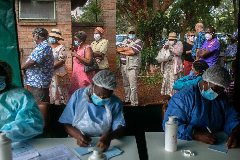 People queue for vaccination outside a tent, while three nurses write down their details on paper.
