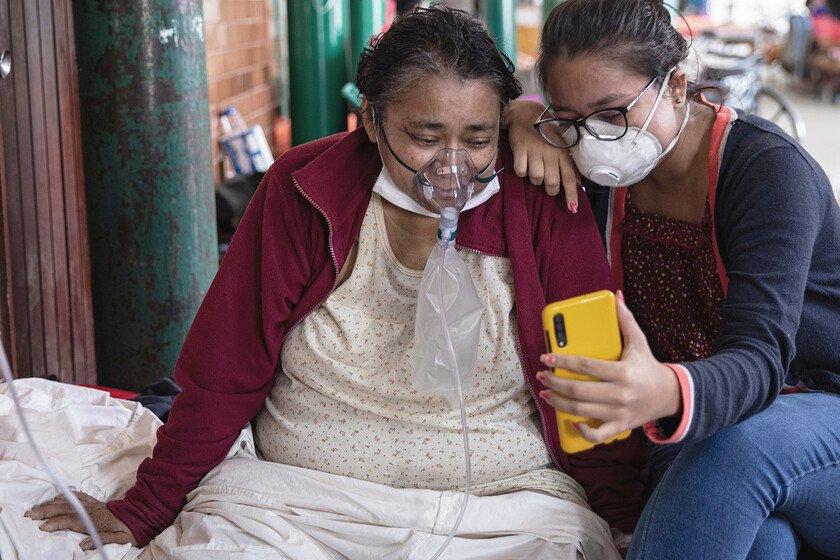 A Covid-19 patient breathing with the assistance of oxygen and her daughter sitting on a bed in a hospital talk to relatives through a mobile phone.