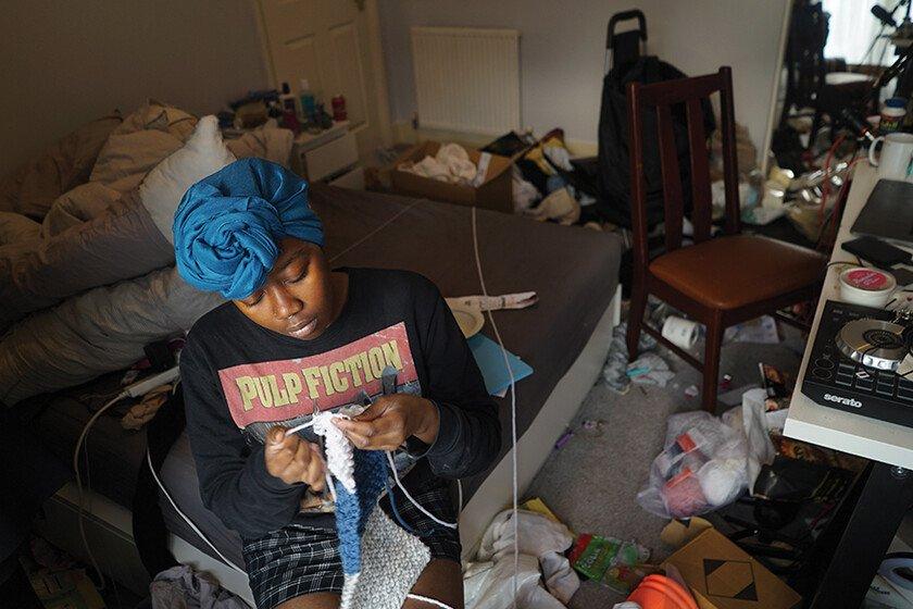 A woman sits on a chair in a cluttered room and knits. 