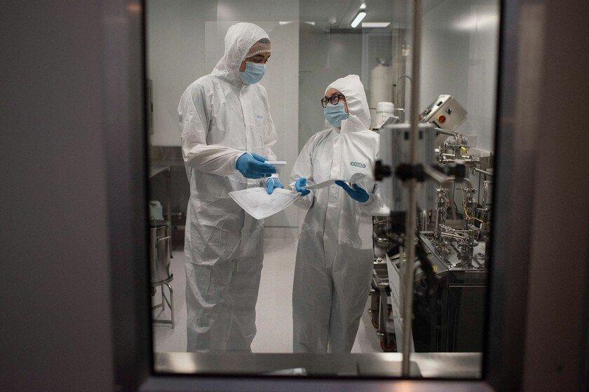 Two scientists in full protective gear stand in a lab discussing lab samples they are holding.