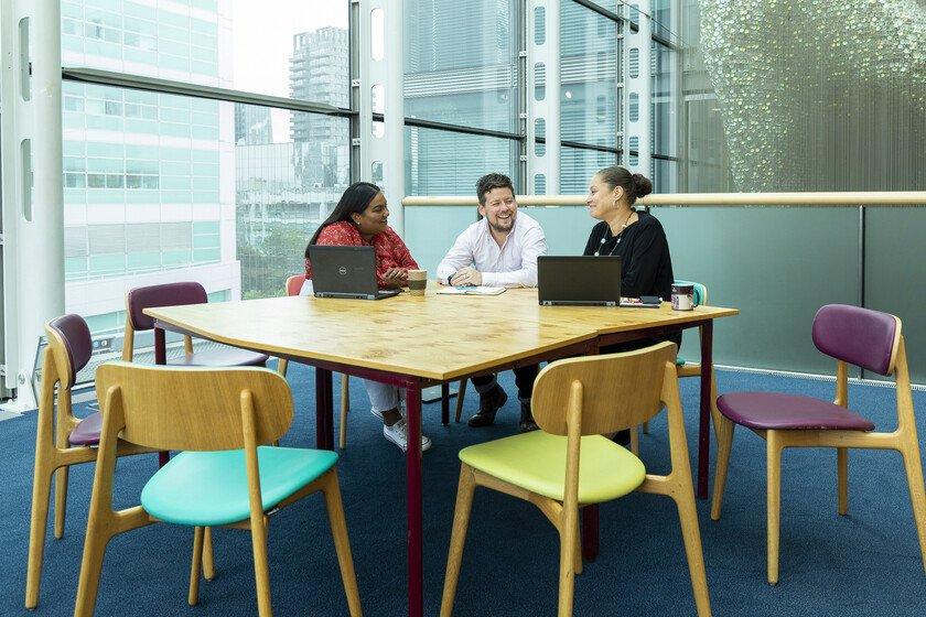 Three Wellcome staff members are sat laughing at a table with laptops. A large window overlooks the city behind them.