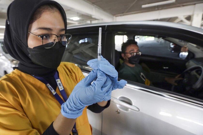 A woman with a face mask holds up a vaccine jab in the foreground while a man with a face mask sits inside a car in the background.