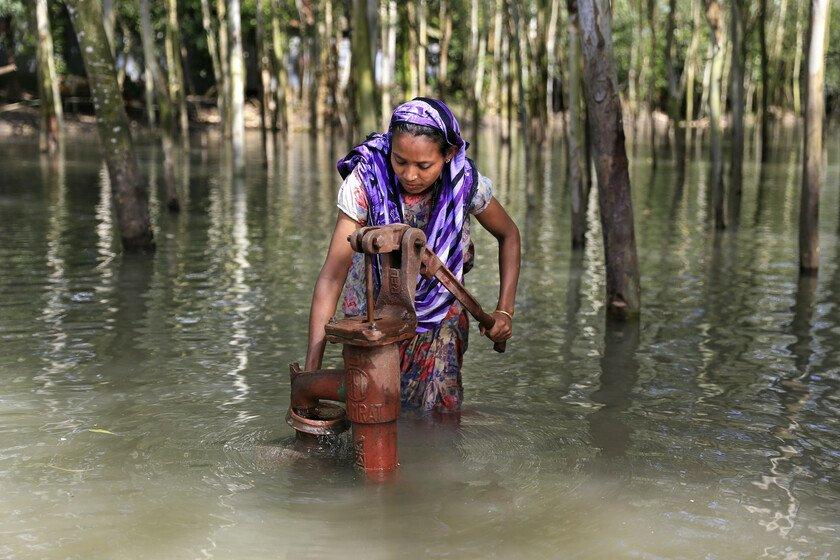 A woman in a purple head scarf stands knee-deep in flood water collecting safe drinking water from a metal hand pump.