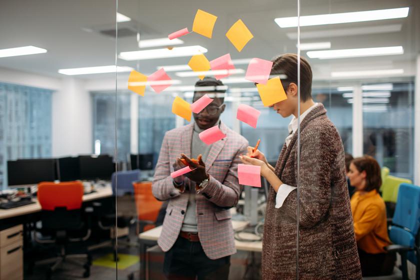 Two people standing in front of a glass wall writing ideas on to sticky note cards.