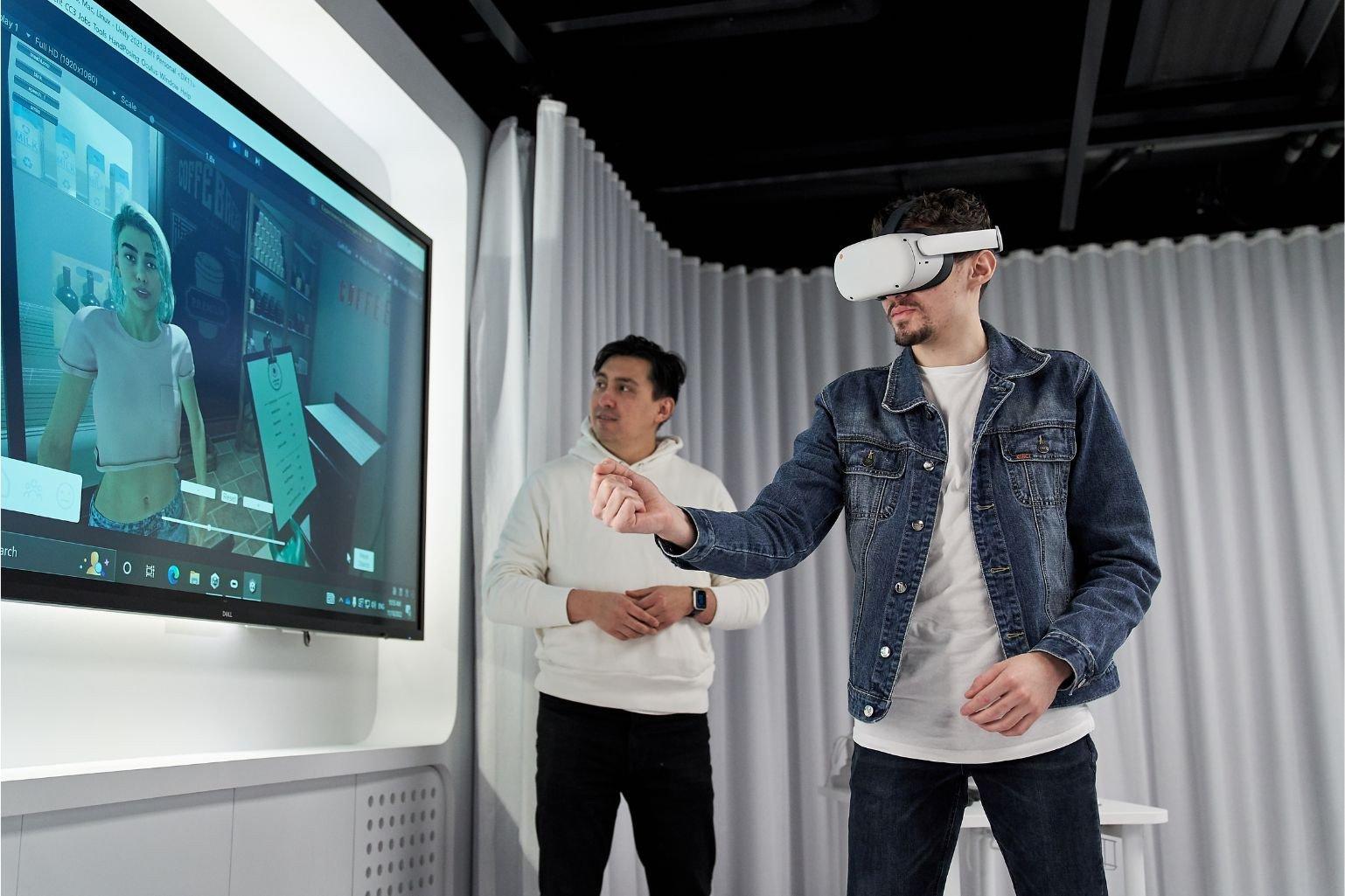 A man wearing a virtual reality (VR) headset standing in front of a TV screen. His hand is outstretched, controlling an animation of a woman on the screen using the VR technology. Another man is standing in the background, watching the screen and the participant's VR control.