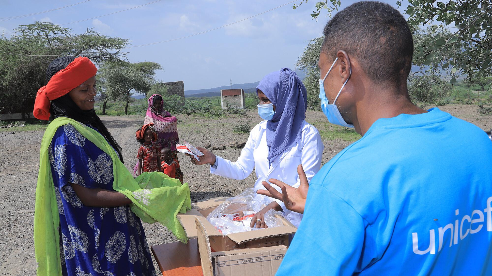Two health workers, one of which wearing a t-shirt with the UNICEF logo, are handing in Ready-to-Use Therapeutic Food to a woman. 