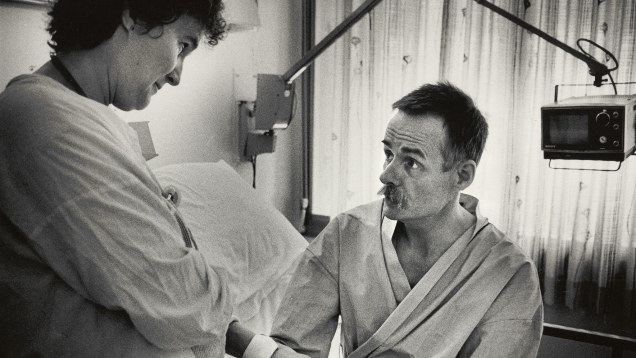 A black and white photo shows a man sat on a bed in a hospital ward. He has a moustache and is wearing a dressing gown as he looks up at a female nurse who is taking his pulse. Behind him the thin curtain is drawn and a small 1980s TV monitor is mounted on a metal arm attached to the wall.