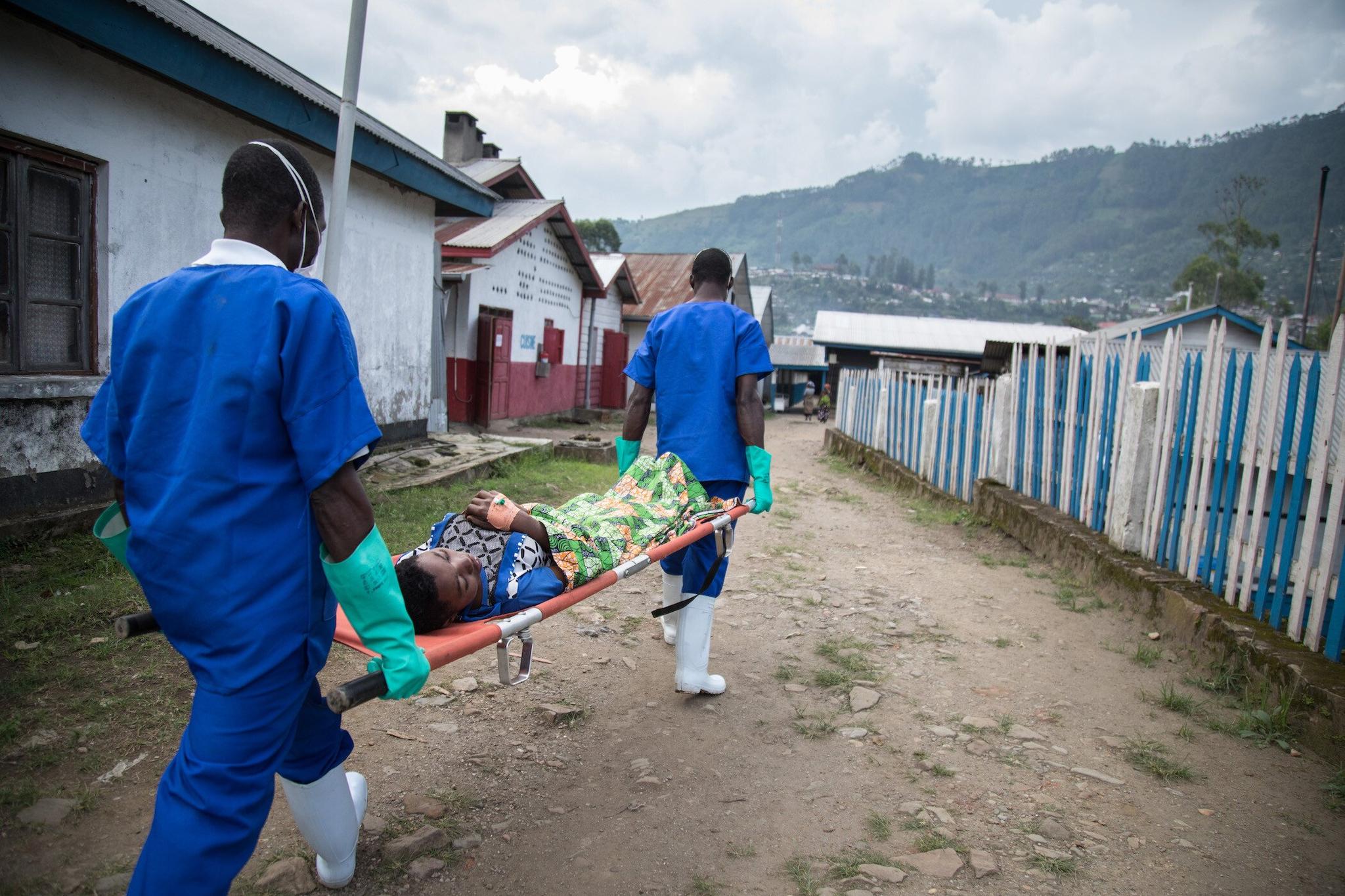 ​​​​​​​Two medical workers in bright blue uniforms carry a patient on a stretcher down a gravel path in a town in the Democratic Republic of the Congo.