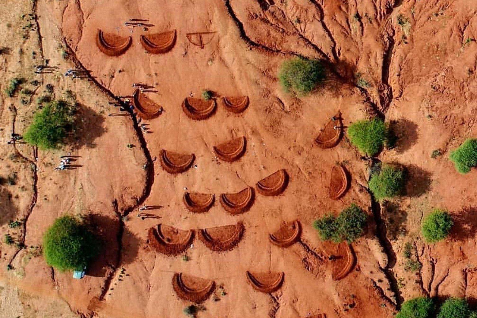 An ariel photo shows a series of large, shallow, half moon-shaped holes being dug into a plain of dry red soil. A few green trees are scattered across the plain, and a small group of people are clustered around a few of the holes.