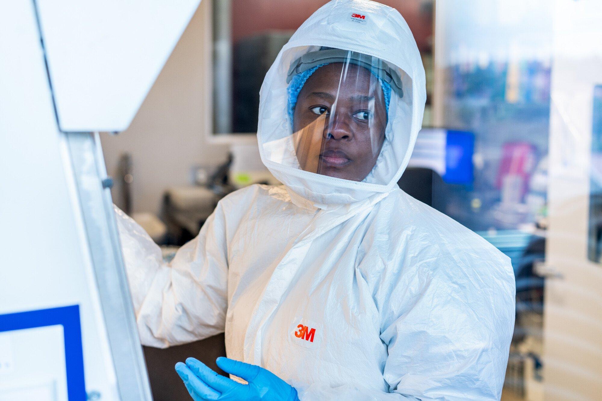 A female researcher in a full body hazmat suit, face mask and gloves is working in a lab.
