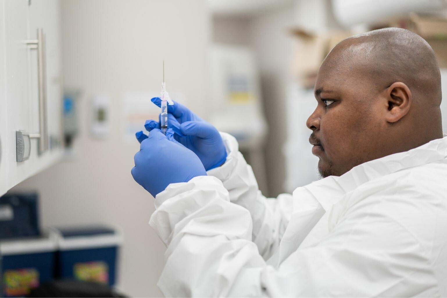 A man wearing a white lab coat and blue gloves is holding and examining a vaccination needle in a laboratory