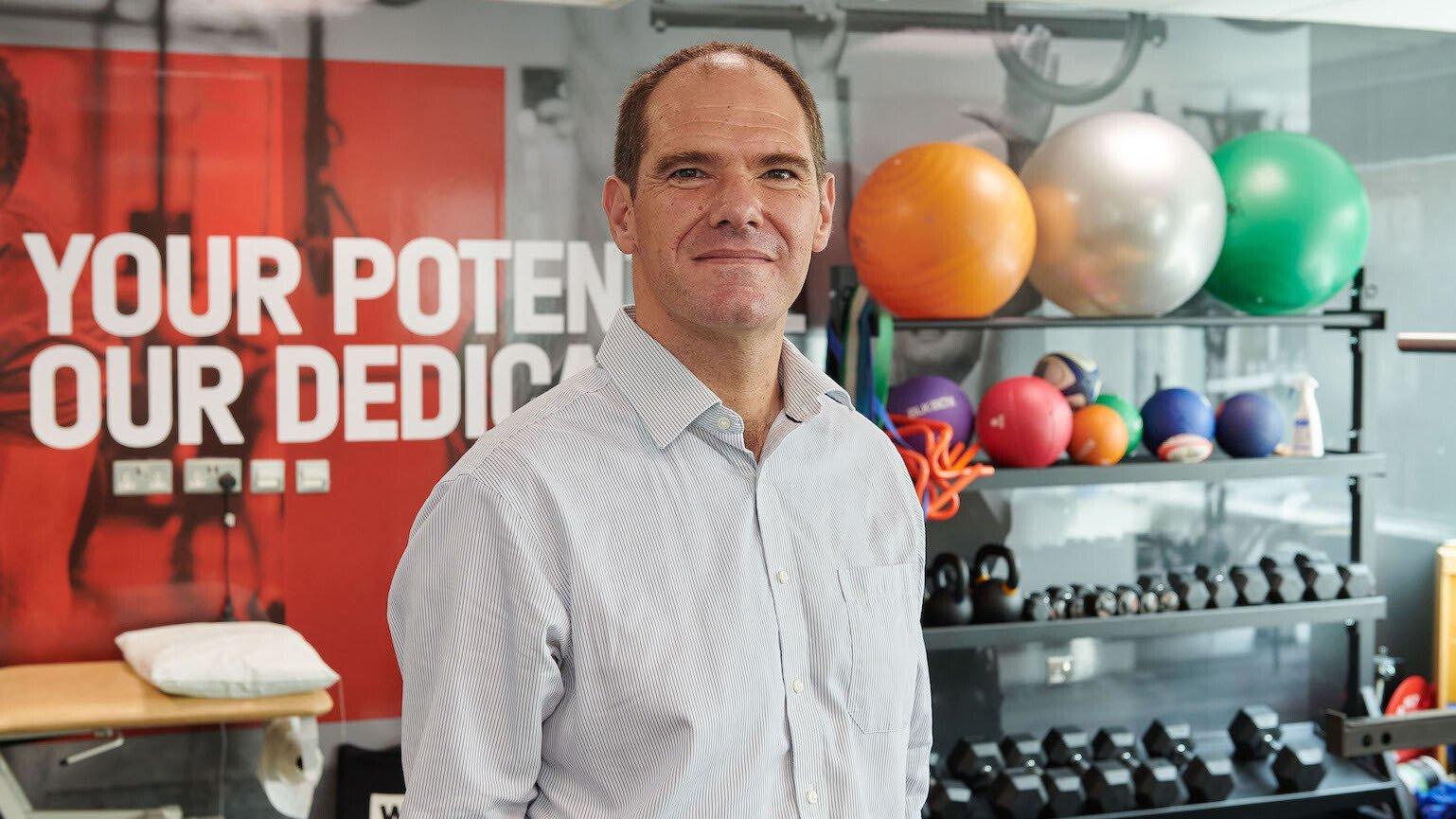 Professor Jonathan Roiser smiles at the camera. He is in a gym, standing in front of a shelf of exercise balls and weights.